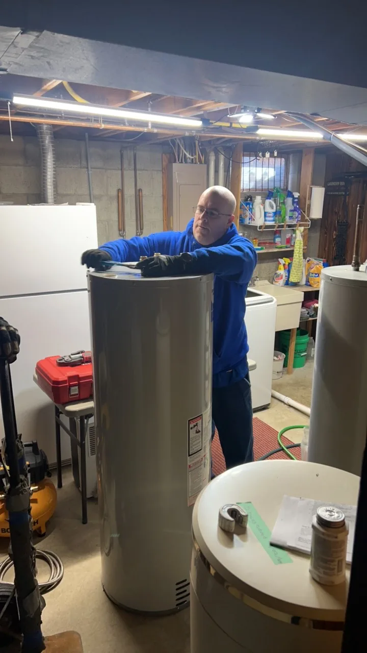 Plumber in blue jacket inspecting a tall cylindrical water heater in a workshop.